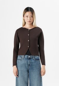 Jonge vrouw met steil blond haar draagt een donkerbruine knopen cardigan over een zwarte top en losse blauwe denim jeans, staande tegen een witte achtergrond.