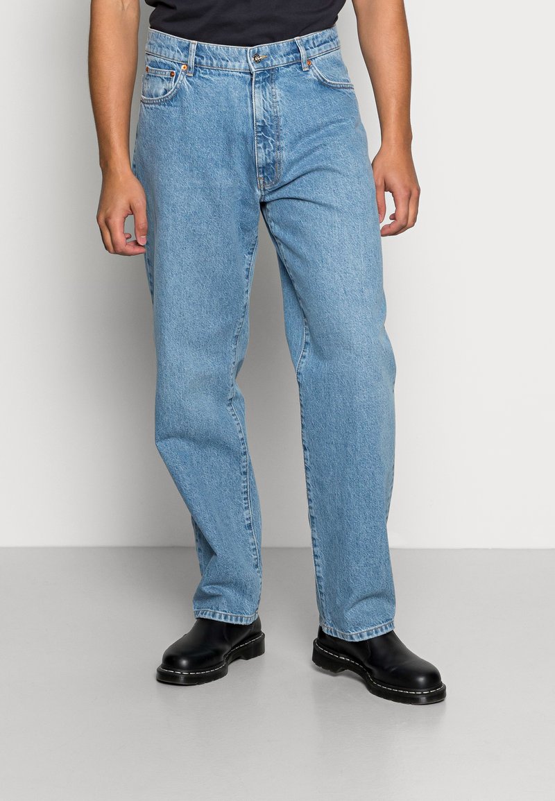 Jeans de denim azul claro com um design de perna reta, cinco bolsos e costura visível. Combinados com botas de tornozelo pretas.