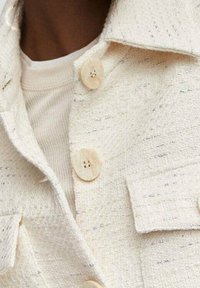 Vaalean kermaisen värinen takki, jossa on teksturoitu kangas ja hopealankoja, kaksi rintataskua ja vaaleat napit. Päällä valkoinen paita.