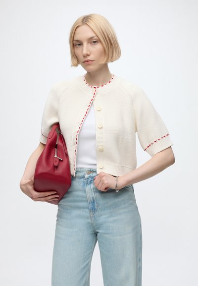 Jeune femme aux cheveux blonds courts porte un cardigan en tricot blanc avec des coutures rouges, un jean bleu clair, et tient un grand sac à main rouge.