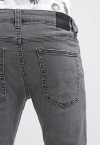 Pantalones vaqueros de mezclilla gris con un corte clásico y una cintura de altura media. Destacan los bolsillos traseros cosidos y una etiqueta de marca de cuero en la cinturilla.