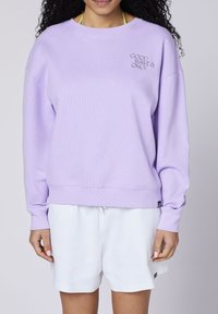 Sudadera de color púrpura claro con tejido texturizado, escote ancho y bordado "SOLO BUENAS OLAS". Combinada con pantalones cortos blancos con cordón.