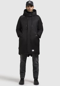 Schwarze Parka mit Kapuze, versehen mit Kordelzügen, Seitentaschen und einem vorderen Druckverschluss. Getragen über eine dunkle Jeans und schwarze Stiefel.