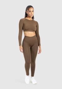 Top corto marrone e leggings a vita alta, entrambi in un tessuto liscio e elasticizzato. Abbinati a sneakers bianche, il modello si trova di fronte a uno sfondo grigio.