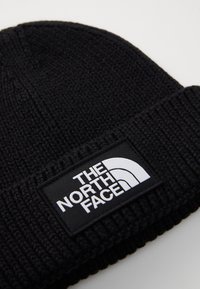Svart stickad mössa med en tjock ribbad textur och en rektangulär vit logotyp med texten "THE NORTH FACE" och grafiskt mönster.