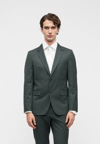 Σκούρο πράσινο κοστούμι με προσαρμοσμένη εφαρμογή, δύο κουμπιά, καμπύλες πέτοι και πλαινές τσέπες. Φορεμένο πάνω από λευκή πουκάμισο με ομαλή υφή.