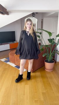 Černé šaty s dlouhým rukávem, výstřihem do V a detailem s vázáním. Modelka má na sobě černé kotníkové boty, stojí na dřevěné podlaze poblíž rostliny.