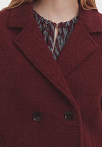 Burgundské dvouřadé sako s texturovanou tkaninou, velkými klopami a černými knoflíky. Pod ním vzorovaná šedá blůza s detaily v podobě klíčové díry.