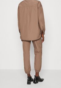 Osoba stojící v volné hnědé košili s dlouhým rukávem a odpovídajícími kalhotami s elastickými manžetami, doplněná černými kotníkovými botami na podpatku.