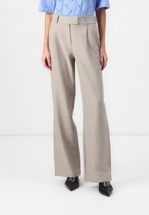 Femme portant un pantalon beige à jambes larges sur mesure, un top en maille bleu clair à manches courtes et des chaussures noires pointues avec boucles argentées.
