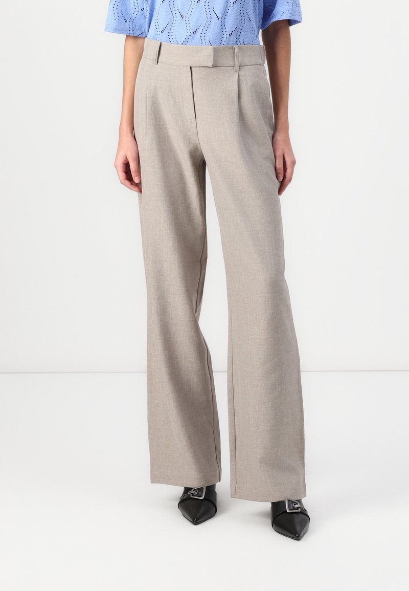 Femme portant un pantalon beige à jambes larges sur mesure, un top en maille bleu clair à manches courtes et des chaussures noires pointues avec boucles argentées.