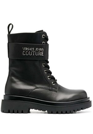 Botín de cuero negro hasta el tobillo con una suela gruesa, que presenta herrajes en plata y "VERSACE JEANS COUTURE" bordado en una correa alrededor de la parte superior.