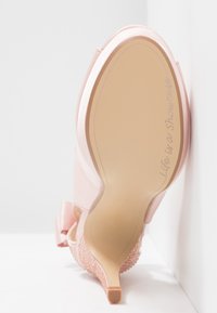 Różowy but na wysokim obcasie z satynowym wykończeniem, teksturowanym obcasem i napisem "Życie jest pokazem" na podeszwie, ustawiony pionowo na białej powierzchni.