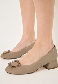 Light taupe suede pumps with a square-shaped plastic accent on the toe and a low block heel, featuring a smooth texture and minimalist design.