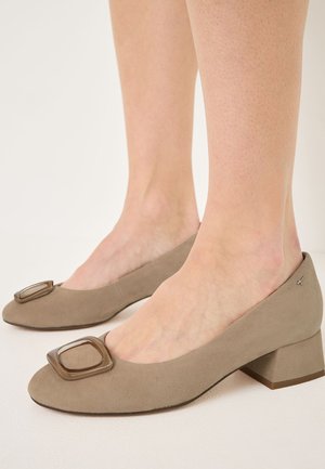 Light taupe suede pumps with a square-shaped plastic accent on the toe and a low block heel, featuring a smooth texture and minimalist design.