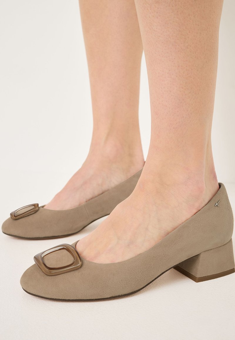 Light taupe suede pumps with a square-shaped plastic accent on the toe and a low block heel, featuring a smooth texture and minimalist design.