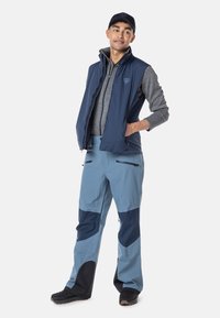 Blaue isolierte Weste über einem grauen Langarmshirt, kombiniert mit hellblauen Schneehosen mit dunklen blauen Akzenten. Schwarze Schuhe.