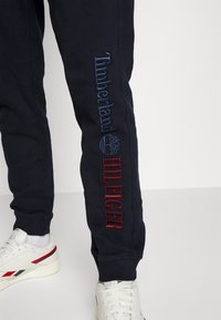 Navy blue sweatpants with embroidered "Timberland" in blue and "Hilfiger" in red on the side, featuring ribbed cuffs and a relaxed fit.