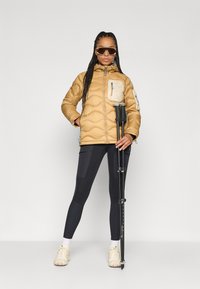 Dikke gouden jas met een crèmekleurige borstzak, zwarte leggings met zijpanelen en beige sneakers; houdende zwarte wandelschoenen.