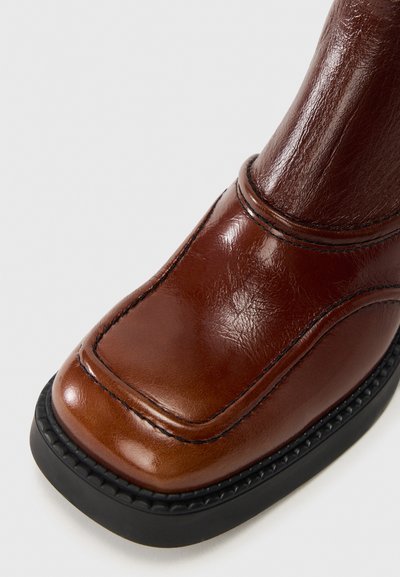 Καφέ δερμάτινο μποτάκι με στρογγυλεμένη μύτη, λεία επιφάνεια, αντίθετη ραφή και μαύρη ελαστική σόλα με υφή στην άκρη.