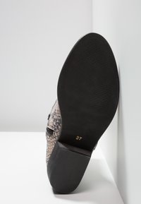 Talons d'une chaussure à motif texturé noir et beige, avec une semelle en caoutchouc présentant des rainures et une étiquette de taille estampillée, placée sur un fond blanc.