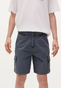 Pantaloni cargo grigi con chiusura a bottone, tasche laterali e una texture leggermente sbiadita. Abbinati a una t-shirt bianca dal taglio largo.