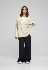 Cremefarbener Strickcardigan mit V-Ausschnitt, sechs großen Knöpfen und langen, oversized Ärmeln, getragen über einem weißen T-Shirt und schwarzen, weit geschnittenen Hosen.