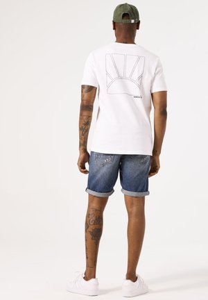 T-shirt bianca a maniche corte con una grafica del sole sulla schiena, abbinata a pantaloni shorts in denim arrotolati e sneakers bianche. Cappellino verde indossato sulla testa.