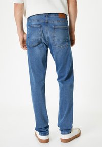 Jeans de mezclilla azul con un diseño de pierna recta, que cuentan con dos bolsillos traseros y un parche de cuero marrón en la cinturilla. Textura suave.