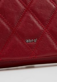Gros plan de cuir matelassé rouge avec couture en zigzag et petit logo métallique "abro" sur un textile ou un accessoire.
