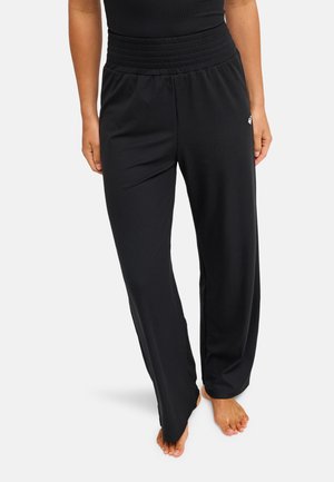 Kobieta w luźnych czarnych, prążkowanych spodniach do relaksu z elastycznym paskiem, stojąca na boso na jednolitym jasnym tle.