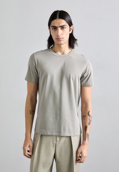 T-shirt in cotone grigio chiaro con colletto tondo, maniche corte e una texture liscia, abbinato a pantaloni sartoriali di colore chiaro.