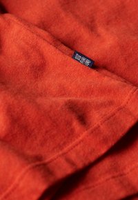 Blødt orange fleece-stof med synlige syninger og en lille mørkeblå mærkat med hvidt stiliseret tekst nær en søm.