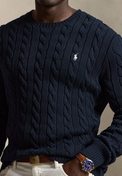 Σκούπε μπλε πουλόβερ με σχέδιο cable-knit και μικρό λευκό κεντημένο λογότυπο, φορεμένο με λευκό παντελόνι και καφέ δερμάτινο ρολόι χειρός.
