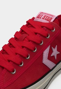 Baskets Converse All Star rouges avec des lacets rouges épais, une étoile rose et un logo chevron, ainsi qu'une semelle en caoutchouc blanche avec une bande noire.