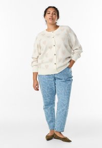 Bēšs kardigans ar sirds rakstiem, ar pogu priekšpusi un ribām pie manžetēm, kopā ar gaisma ziliem rakstainiem džinsiem un olīvzaļām dzīvoklēm.