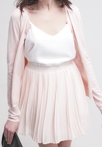 Cárdigan rosa sobre un camisón de satén blanco, combinado con una falda plisada en tono rubor. El conjunto presenta texturas suaves y un ajuste relajado.