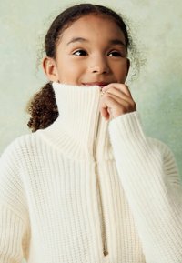 Pull en tricot de couleur crème avec un col haut en côte et un détail de fermeture éclair doré, présentant un motif texturé et des manches amples.