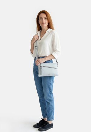 Frau mit roten Haaren, die eine weiße Bluse, blaue Jeans und schwarze Turnschuhe trägt, hält eine hellgraue Umhängetasche mit Reißverschlusstasche.