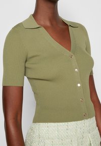 Oliwkowy, prążkowany kardigan z krótkim rękawem, z dekoltem w kształcie litery V i pięcioma złotymi guzikami z przodu. Teksturowany materiał, dopasowany krój.