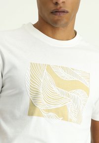 La camiseta de manga corta blanca presenta un gráfico texturizado en amarillento pálido y blanco, que representa patrones de hojas curvadas sobre un fondo cuadrado.
