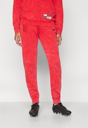 Personne portant un pantalon de survêtement rouge Liverpool FC Nike et un sweat-shirt à capuche assorti, avec des crampons de football noirs sur un sol et un fond gris clair.