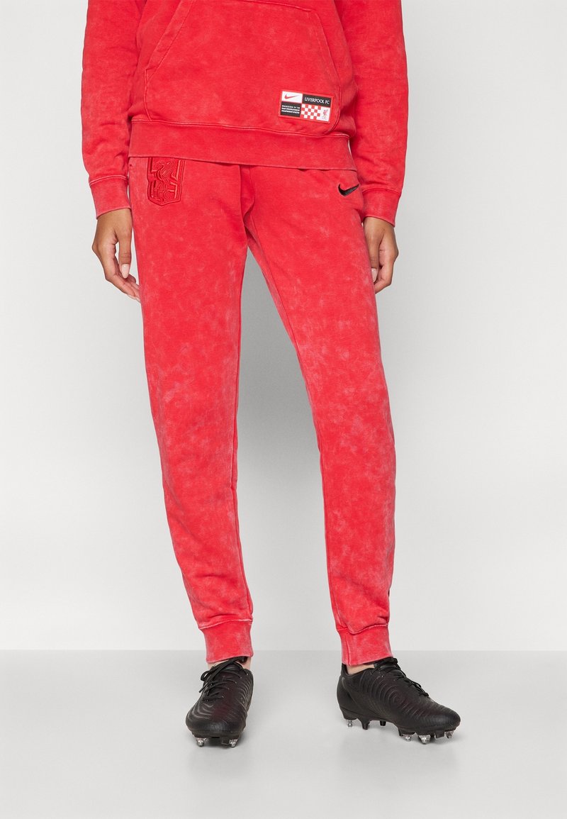 Osoba ubrana w czerwone spodnie dresowe Liverpool FC Nike i pasującą bluzę z kapturem, na szarym jasnym tle, nosząca czarne korki piłkarskie.