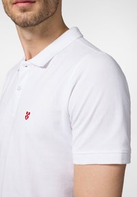 Biała koszulka polo wykonana z bawełny, z kołnierzykiem, krótkimi rękawami i małym czerwonym logo na piersi. Teksturowana tkanina.