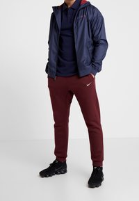 Marineblauwe lichtgewicht jas met rode capuchon, marineblauwe polo, kastanjebruine sweatpants met het Nike-logo en zwarte sportschoenen. Textuurmaterialen zichtbaar.