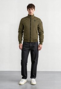 Giacca bomber verde oliva con colletto alto, zip frontale, tasche laterali e polsini e orlo a costine. Abbinata a pantaloni neri e sneakers bianche.