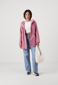 Ružová vodoodolná bunda s kapucňou, dvoma prednými vreckami a patentkami, oblečená cez biele crop top a roztrhnuté modré džínsy, drží bielu tašku.