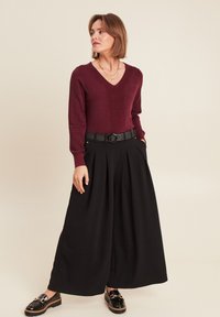 Bordeaux V-hals trui met een gestructureerd middenpaneel, gecombineerd met hooggetailleerde zwarte wijde broek en glanzende zwarte loafers met een gouden gesp.