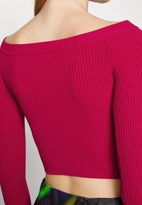 Top corto ajustado de canalé en rosa brillante, con un diseño de hombros al descubierto y mangas largas. La tela presenta rayas verticales con una textura suave.