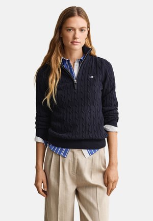 Kvinde iført navyblå kabelstrikket halv-lynlås sweater over blå og hvide stribede skjorte og beige plisserede bukser, stående med afslappet kropsholdning.